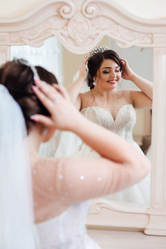 Beautiful bride in white dress near a mirror. Bride straightens the crown. Beautiful happy bride preparing for her wedding. Bride with elegant hairstyle and makeup. Beauty woman