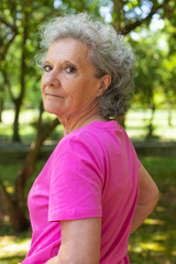 Serious confident old lady walking in park. Side and back of senior grey haired woman in casual turning face to camera. Senior woman outdoors concept