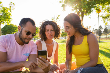 Focused Latin guy showing content on phone to female friends. Man and women sitting on grass in park, watching and discussing video on smartphone. Communication outdoors concept