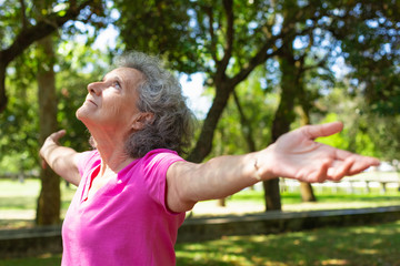 Peaceful tranquil old lady relaxing and exercising in park. Senior grey haired woman in casual standing in park, opening hands and looking up. Freedom concept