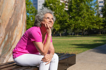 Dreamy old lady relaxing in city park. Pensive senior grey haired woman in casual sitting on bench outdoors, leaning chin on hand, thinking, looking up. Thinking in park concept