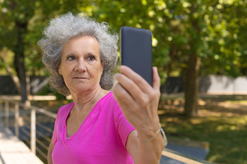 Obraz premium Serious focused old lady with smartphone having video call. Senior grey haired woman in casual talking on mobile phone while walking in park. Communication concept