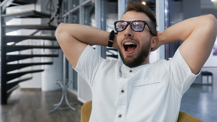 Young business man with a shocked expression working in cozy creative office. Man with beard in formal clothes and eyeglasses, confident and successful  celebrating a triumph with arms up