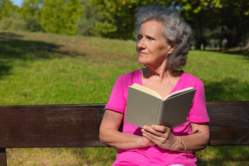 Frowning old lady with book staring away. Senior grey haired woman in casual sitting on park bench, holding book and looking into distance. Reading book outdoors concept