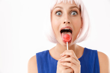 Portrait of young surprised woman wearing pink wig licking lollipop at camera