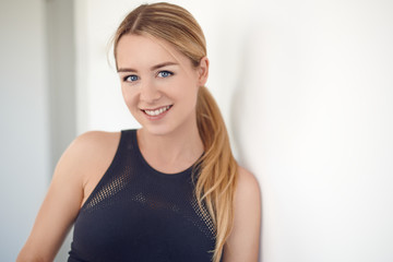 Pretty young blond woman with a friendly smile and her hair in a ponytail leaning on an interior...