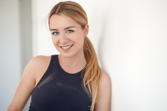 Pretty young blond woman with a friendly smile and her hair in a ponytail leaning on an interior white wall looking at the camera in a head and shoulders closeup portrait