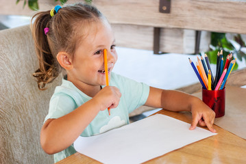 Little girl smiles, sits at a table, summer playground cafe with colored pencils in hand.