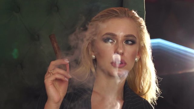 A luxurious young girl in a strict jacket sits in a chair in an expensive restaurant and smokes a cigar, she lets out a lot of smoke. Slow motion.