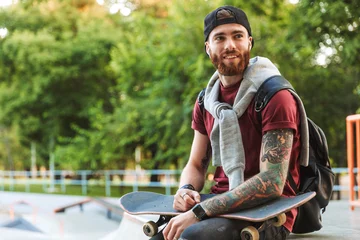 Fotobehang Skateboard Attractive cheerful young man sitting at the skate park ramp  © Drobot Dean