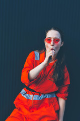 Young woman in red overalls and red sunglasses posing near black wall with candy