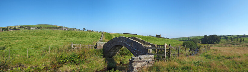 Fototapeta na wymiar a panoramic view of an ancient stone packhorse bridge crossing a stream in west Yorkshire dales countryside with the village of colden in the background