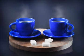 Two cups of fresh coffee on a wooden stand. Steam from a coffee mug. Blue mugs with a hot drink on a dark background next to the sugar