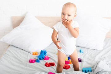 Naklejka premium Smiled barefoot baby in white clothes standing on bed with toys and taking fingers into his mouth