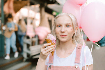 Portrait of a funny cute girl with blond hair holds a pink ice cream cones and balloons, looks emotionally into the camera and smiles. Cute blonde lady with makeup eating ice cream on a walk.