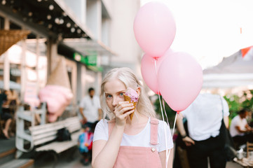 Street portrait of a funny blonde girl standing on the street with ice cream and pink balloons in her hands and looking into the camera with a serious face, wearing a pink sundress and a white T-shirt