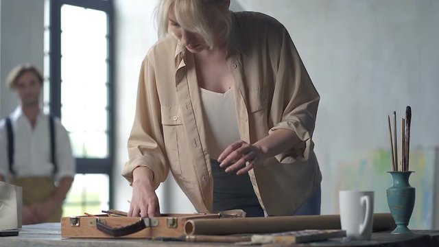 Female painter choosing painting tools at the table, while mannequine man is sitting on the chair and waiting at the background. Slowmotion.