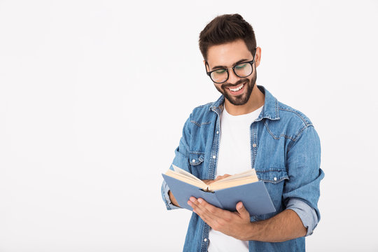 Image of young happy man wearing eyeglasses reading book and laughing