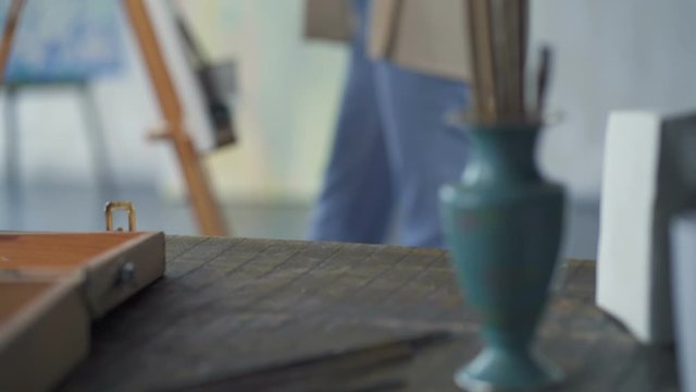 Handheld footage of artist woman painting. Focus on the table with painting tools, then ath the woman. Slowmotion.