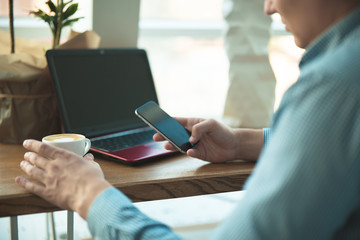 young handsome businessman checks his smartphone holding cup of coffee while working in cafe near office