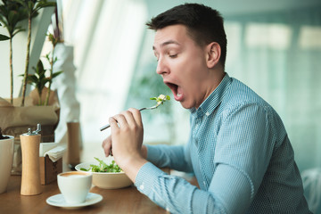 young handsome man puts salad in his mouth and drinks coffee for lunch during break at cafe near office looks hungry