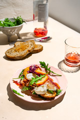 Parma ham, mozzarella and peaches sandwich on a plate, bowl of arugula, some bread and rose wine shot with hard light
