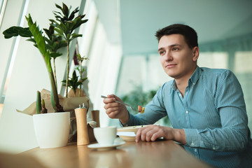 young handsome man holding fork in his hand eats salad and drinks coffee for lunch during break at cafe near office looks hungry