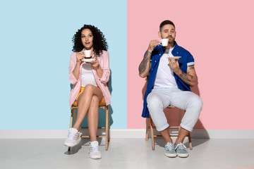 Fototapeta premium Young emotional caucasian couple in bright casual clothes posing on pink and blue background. Concept of human emotions, facial expession, relations, ad. Woman and man drink coffee or tea and smile.