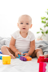 Funny barefoot baby in white clothes sitting on bed, playing with colorful construction and looking at camera