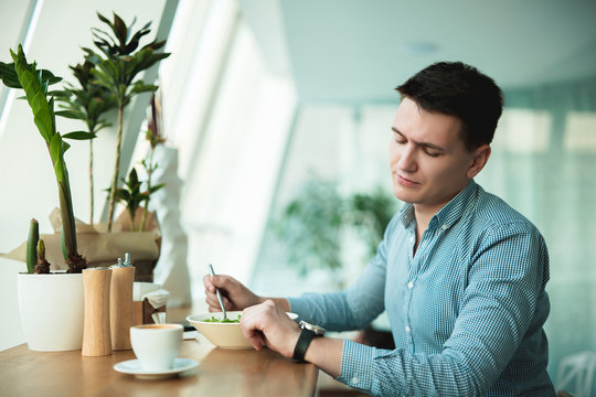 young handsome man looks at his wristwatch while eating salad and drinking coffee during break at cafe waiting for his patner to come