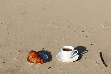 A cup of coffee with a croissant stands on the seashore
