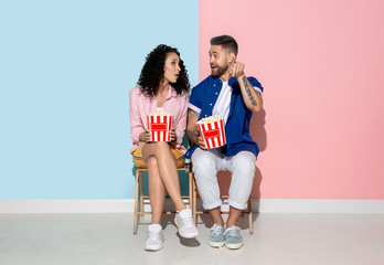 Young emotional caucasian couple in bright casual clothes posing on pink and blue background. Concept of human emotions, facial expession, relations, ad. Woman and man watching TV with popcorn.