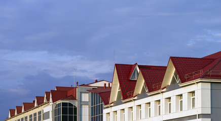 The roof of a building made of red metal tile with ventilation pipes and lightning rods against a blue sky.