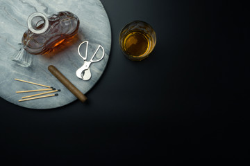 top view of a Cuban cigar and a stainless steel cigar cutter, a whiskey decanter with matches on the marble tray, a glass of bourbon whiskey on the black table