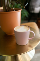 Coffee latte in pink mug sitting on gold table next to plant, hipster coffee shop, close up