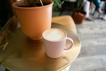 Coffee latte in pink mug sitting on gold table next to plant, hipster coffee shop, close up