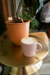 Coffee latte in pink mug sitting on gold table next to plant, hipster coffee shop, close up