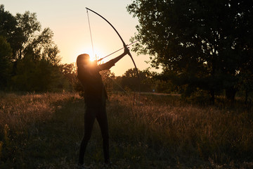 A woman shoots a bow in nature at sunset.
