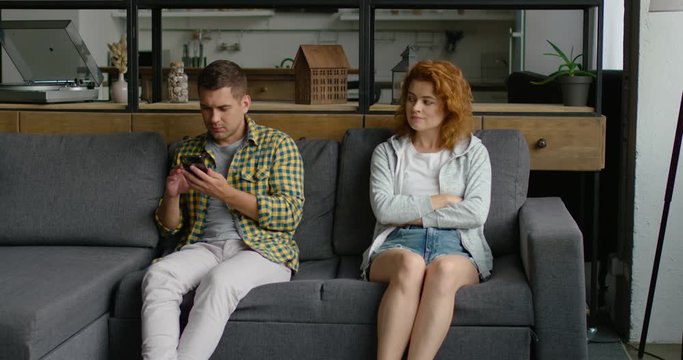 Man is sitting on sofa, using his smartphone, gadget, relaxed and in good mood, woman is angy and annoyed, sitting next to him, getting nervous by his behavior, indoor at home. 4K, shot on RED camera.