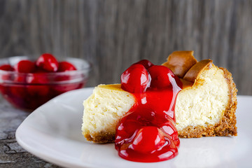 A slice of New York style cheesecake with cherry sauce on top.  Bowl of cherry sauce blurred in the background.  White plate.  Neutral background.