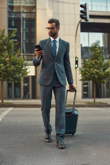 Always in touch. Full length of young and handsome bearded man in suit pulling his luggage and looking at his smartphone while walking outdoors