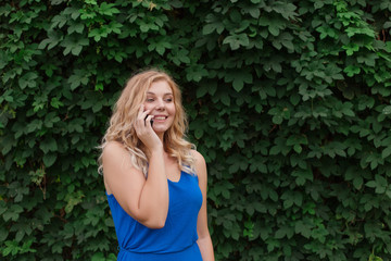 Beautiful young girl in a blue dress talking on the phone. Against the background of wild grapes, summer day. Copy space.