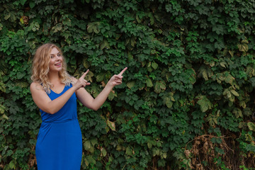 Young beautiful girl on a background of a wall of wild grapes, points fingers to the side. Copy space.
