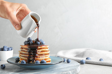 Woman pouring chocolate syrup onto fresh pancakes with blueberries at grey table, closeup. Space for text - obrazy, fototapety, plakaty