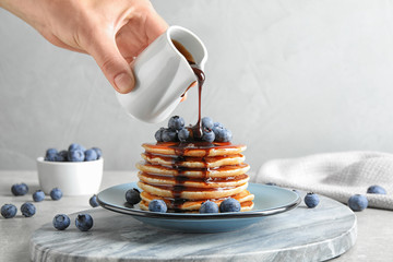 Woman pouring chocolate syrup onto fresh pancakes with blueberries at grey table, closeup - obrazy, fototapety, plakaty