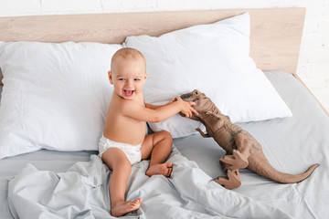 Happy little child sitting on bed, smile and playing with brown toy dinosaur