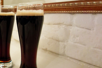 Two glasses poured with dark beer or soft drink with foam head. Close up image against white brick wall in bar or pub