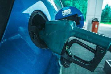 A gasoline filling gun is inserted into the tank of a blue car at a gas station.