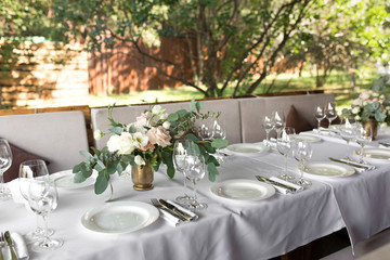 Wedding table setting decorated with fresh flowers in a brass vase. Wedding floristry. Banquet table for guests outdoors with a view of green nature. Bouquet with roses, eustoma and eucalyptus leaves