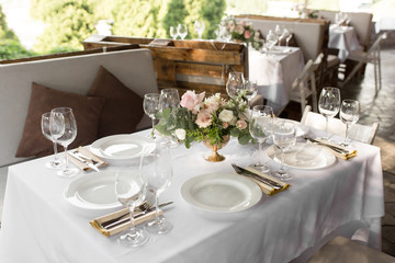 Wedding table setting decorated with fresh flowers in a brass vase. Wedding floristry. Banquet table for guests outdoors with a view of green nature. Bouquet with roses, eustoma and eucalyptus leaves
